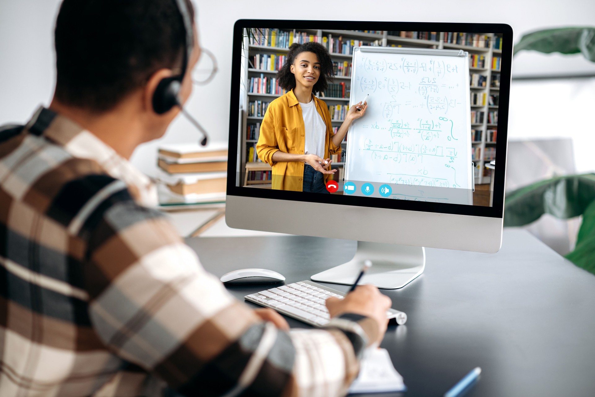 E-learning concept, webinar, online lecture. Over shoulder view of male student on a computer screen, female friendly african american teacher on the screen conducts an online lesson,a guy takes notes