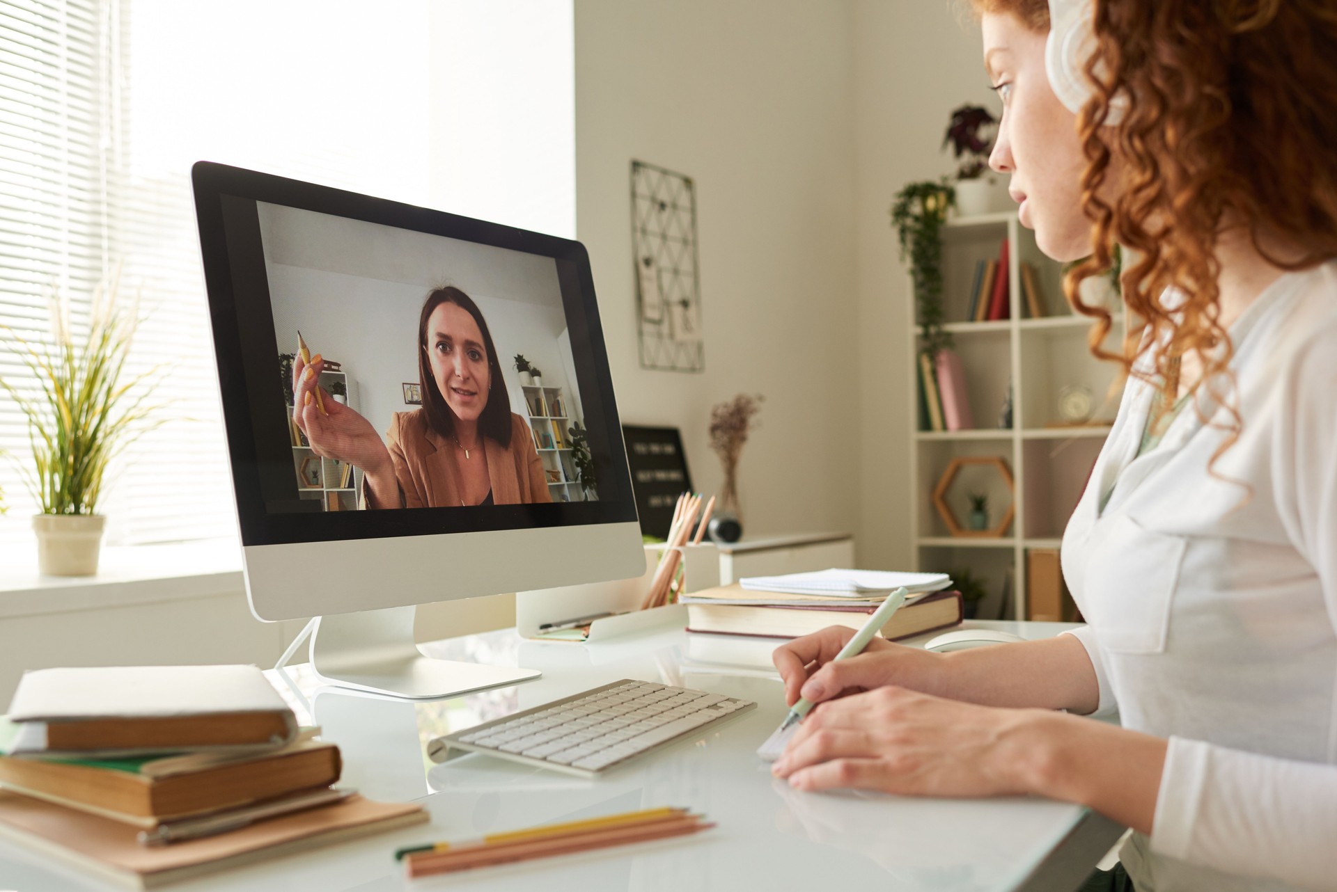 Online tutor assisting student girl with coursework during video conference, student girl with curly hair making notes